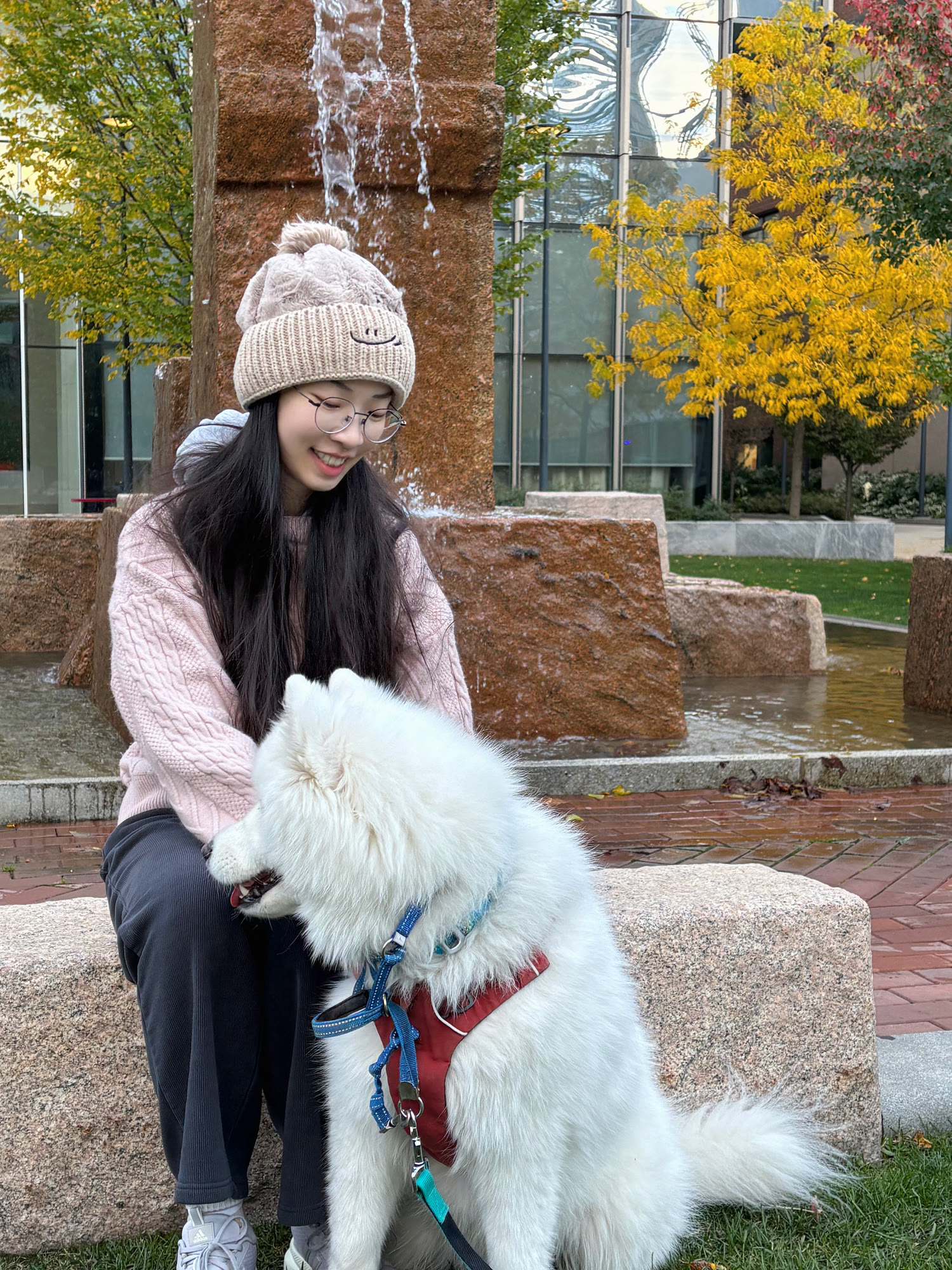 Jiawen Wu smiling while spending time outdoors with her large white dog beside a fountain in autumn.