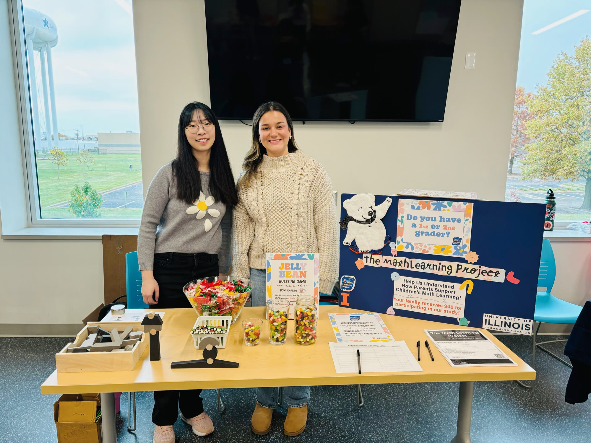 Jiawen Wu and her research assistant Alaina standing behind hands-on mathematics activities and a Math Learning Project information display at a community STEM event.